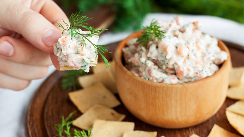 A hand holding cracker with scooped shrimp dip and bowl in background