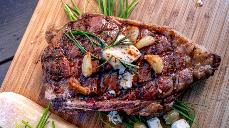Grilled ribeye steak topped with butter, garlic, and herbs on a wooden cutting board