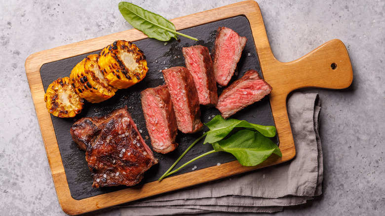 Slices of New York strip steak on a wooden cutting board with fresh arugula and grilled corn