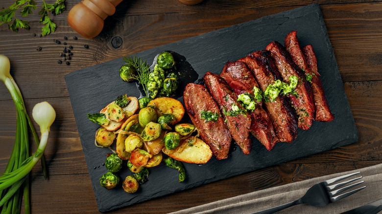 Sliced grilled hanger steak on black slate with roasted potatoes and brussels sprouts