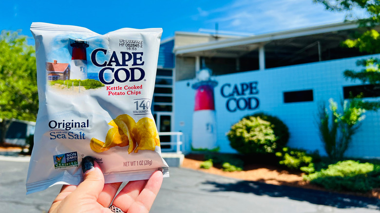Hand holding small bag of Cape Cod Original Sea Salt Potato Chips in front of building with matching logo