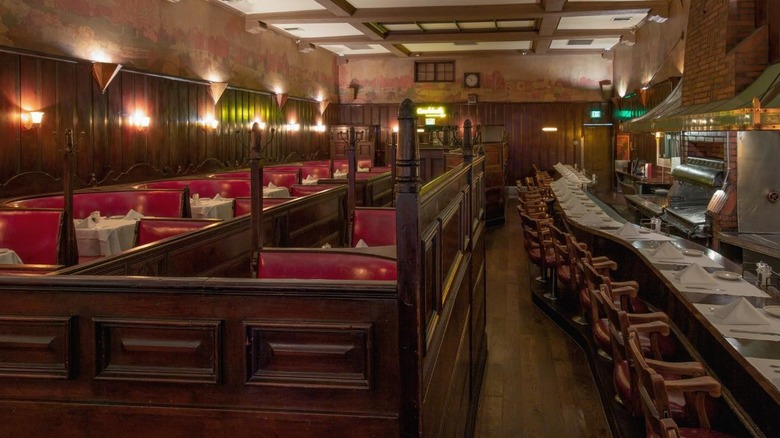 Musso & Frank dining room with circular red leather booths on the left and 2-top red leather booths on the right, lit by warm yellow sconces, bar seating on the right,