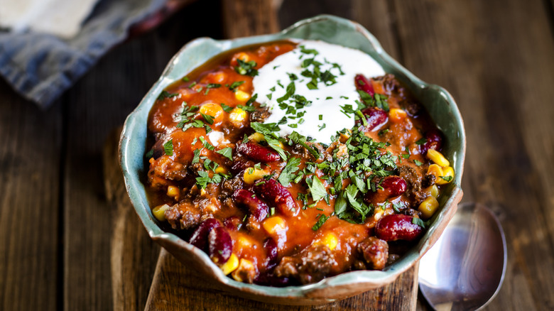 A clay bowl of chili with sour cream and herbs on top on a wooden table