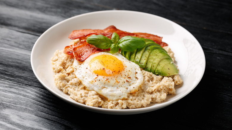white bowl with oatmeal, bacon, avocado, and fried egg on a wooden table