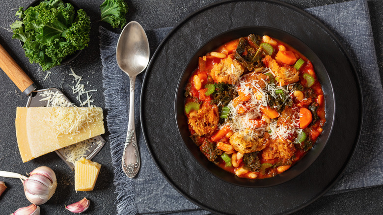 Black bowl of ribollita surrounded by fresh kale, garlic, and parmesan cheese