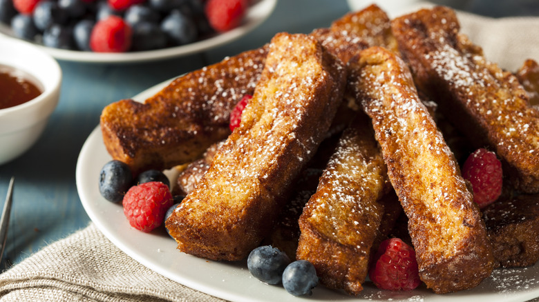 White plate of French toast sticks garnished with powdered sugar and fresh berries