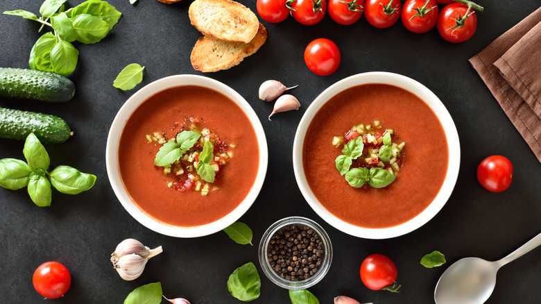 Two white bowls of gazpacho soup surrounded by its ingredients