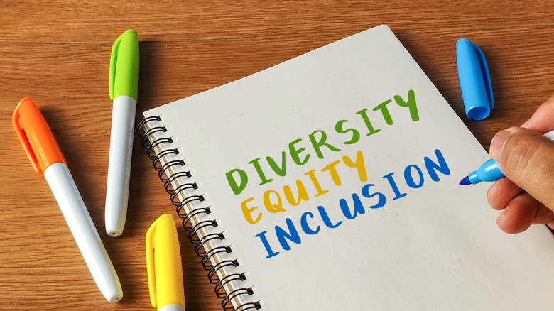A hand holding a marker over a notebook with "Diversity Equity Inclusion" written on it in different colors
