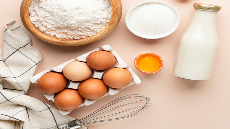 Various sized bowls of flour, butter, sugar, and cinnamon and a glass bottle of milk on a light pink background
