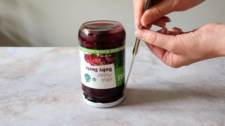 A woman's hands are shown using a butter knife to break the seal of a jar.