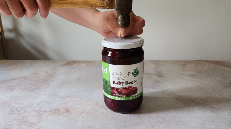 A woman's hands are shown using a hammer and nail to break the seal on a jar.
