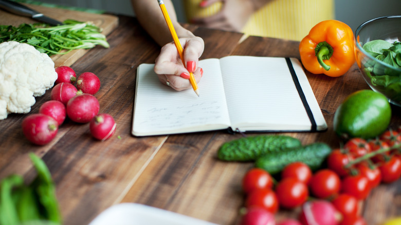 Hand writing a list in a notebook on a wooden surface with fresh vegetables surrounding