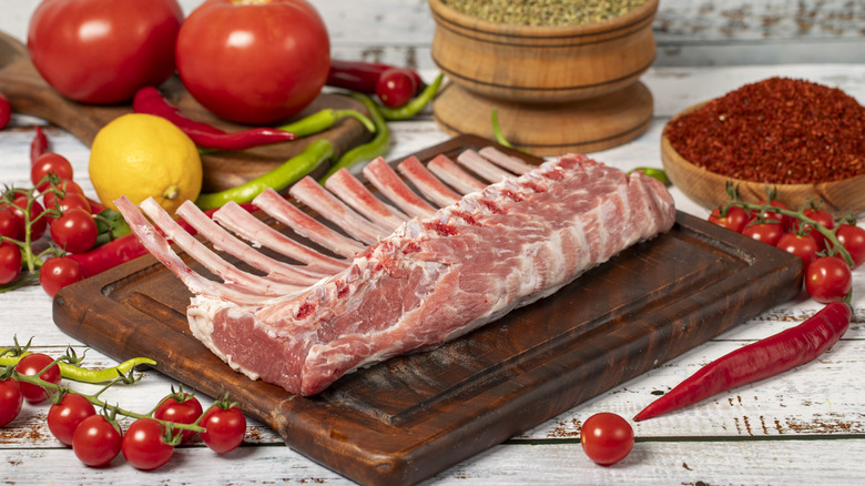 A rack of lamb on a cutting board with spices and tomatoes in the background