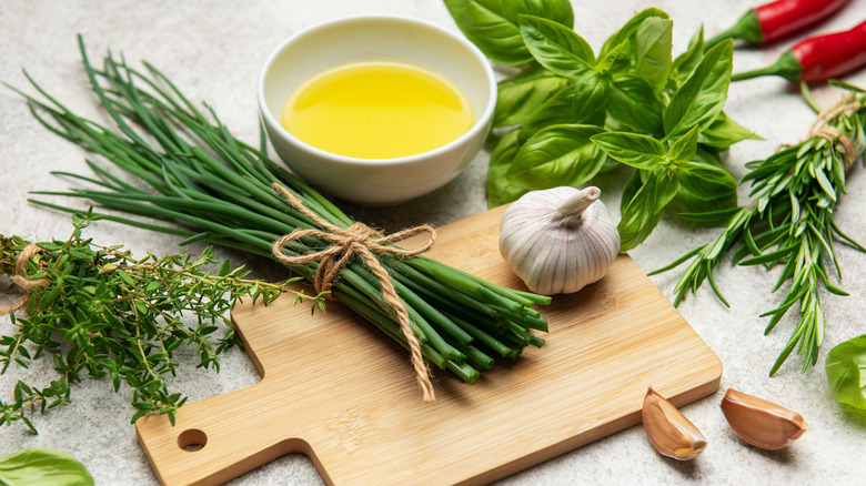 A dish of olive oil with garlic and fresh herbs surrounding it next to a wooden cutting board
