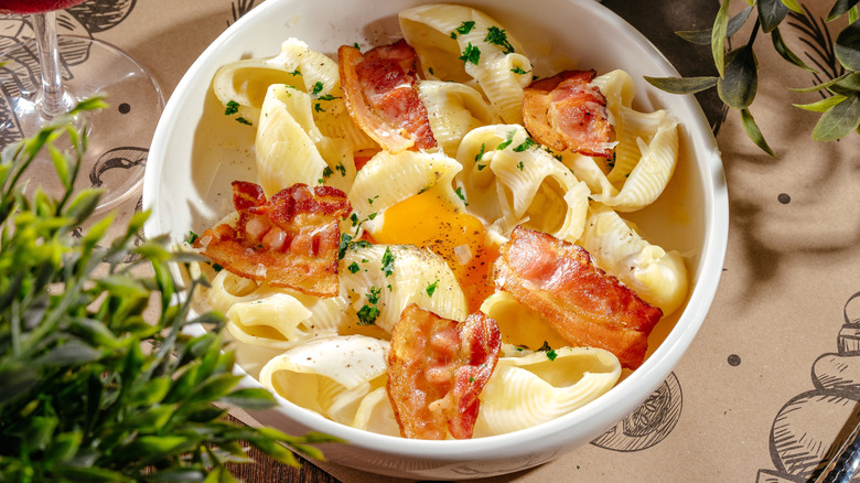 Large shell pasta with bacon pieces in a white bowl with greens on the sides