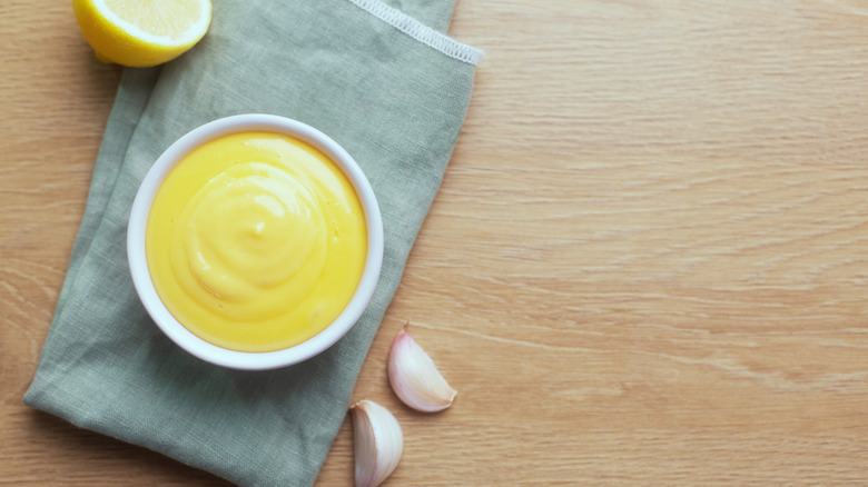 Aioli in a white ramekin placed on a grey cloth napkin with raw garlic and a lemon wedge