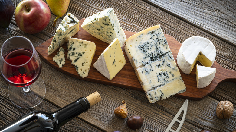 Assorted blue cheeses on a wooden board next to fresh fruit and red wine