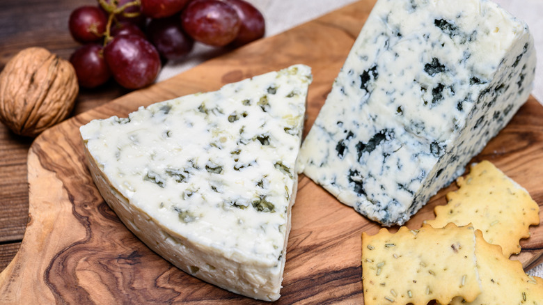 Wedges of Danish blue cheese on a wooden board next to crackers, grapes, and walnuts