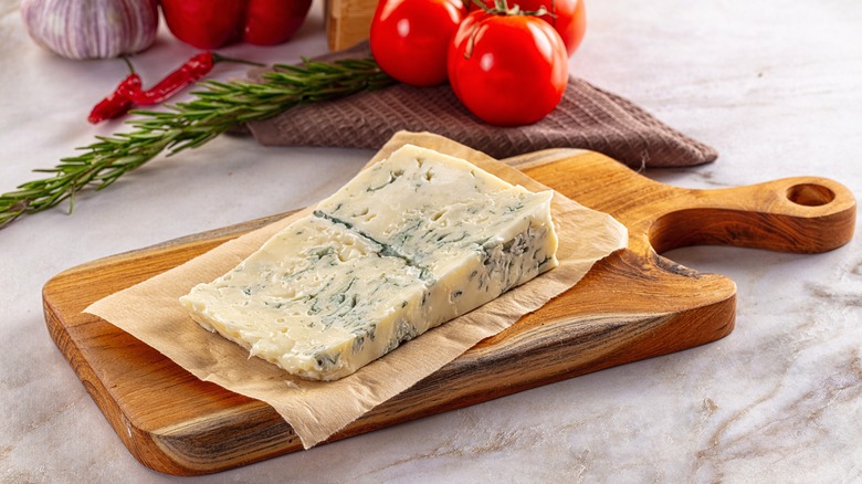 A wedge of Gorgonzola on paper on a small wooden board in front of tomatoes, peppers, and rosemary