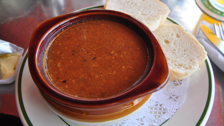 A crock filled with red chowder on a green and white plate with a doily, and two slices of bread alongside