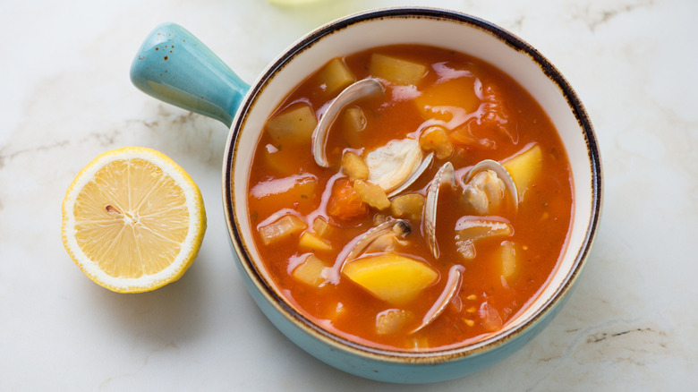 Turquoise serving bowl of Manhattan Clam Chowder on white marble bg with half a lemon alongside