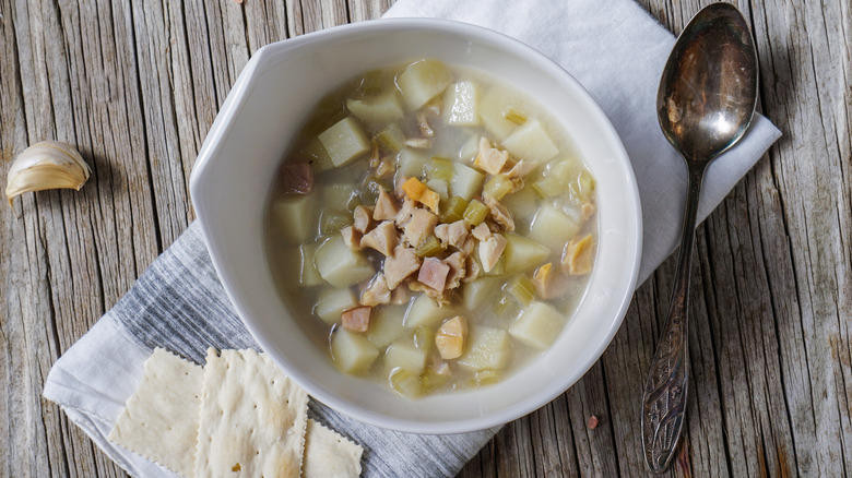 White bowl of Rhode Island Clam Chowder on wood background with serving spoon, napkin, saltines, and garlic clove