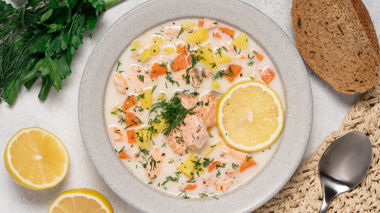 Large white bowl of salmon chowder with lemon and dill garnish, herbs, lemon, brown bread, and spoon on the surface surrounding it