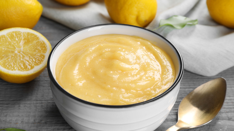 Delicious lemon curd in bowl, fresh citrus fruits and spoon on grey wooden table, closeup
