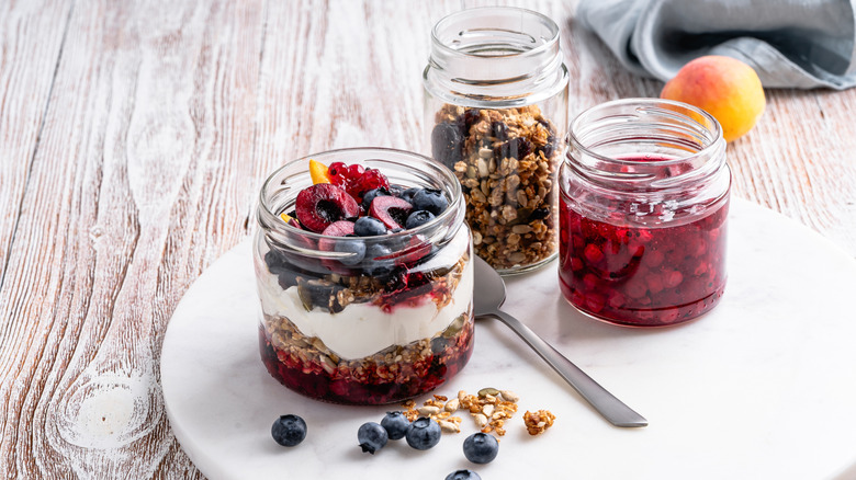 Breakfast with yogurt, granola and berries and berry jam preserve in glass jar, on white wood background
