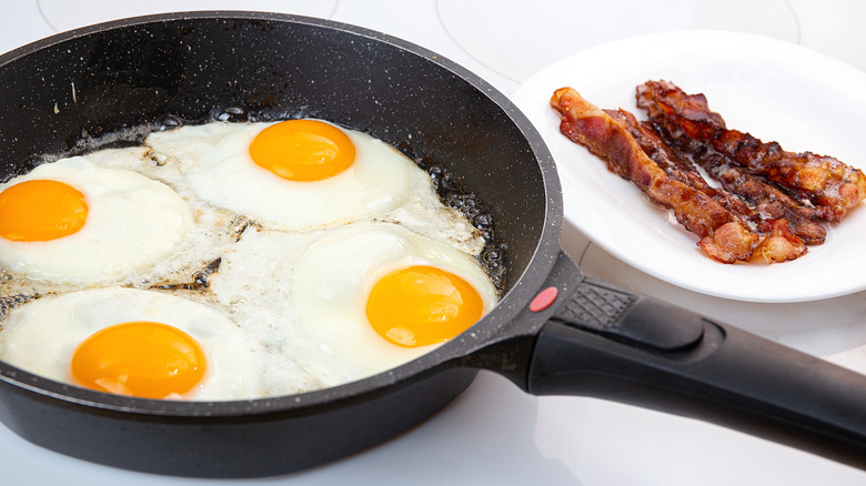 Four eggs frying in a skillet next to a plate of bacon