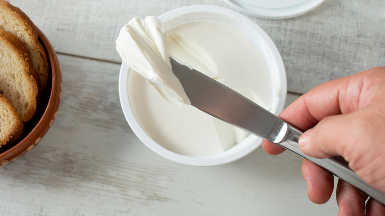Hand holding a butter knife, taking a generous portion of cream cheese from a container