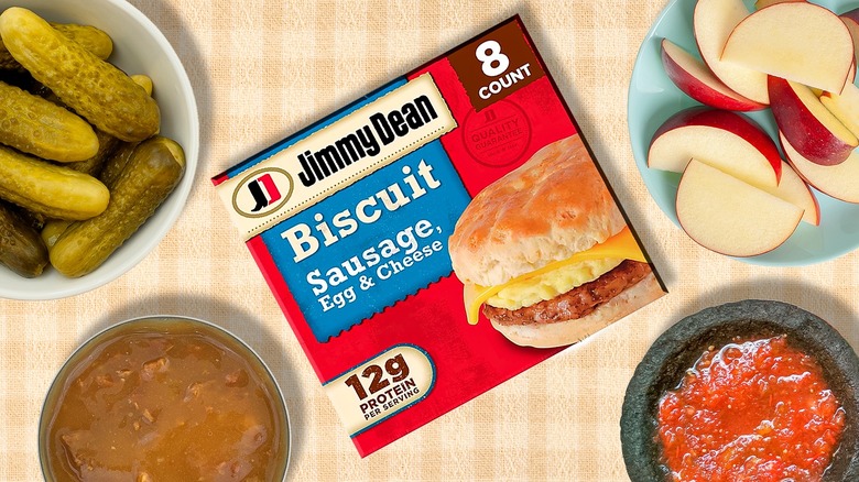 A box of Jimmy Dean breakfast sandwiches surrounded by dishes and bowls of upgrade options like pickles, apple slices, and salsa