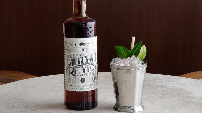 A bottle of Ancho Reyes chile liqueur next to a cocktail in a copper mug garnished with mint and lime