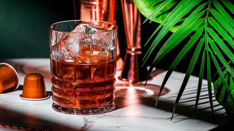 A coffee cocktail in a short rocks glass on a bar next to a plant and two espresso pods
