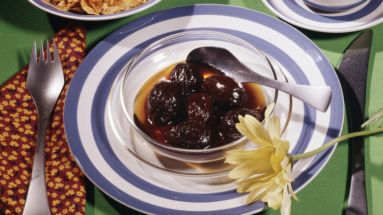 A glass bowl of stewed prunes on a plate laid out on a breakfast table next to a yellow flower