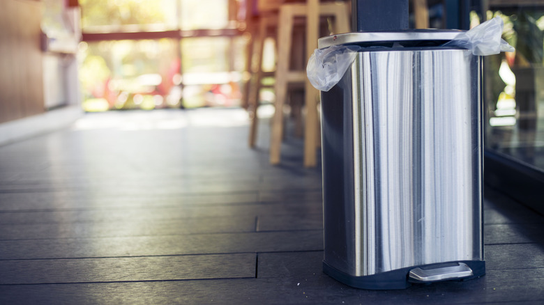 A metal trash can with a lid sits on a gray hardwood floor.
