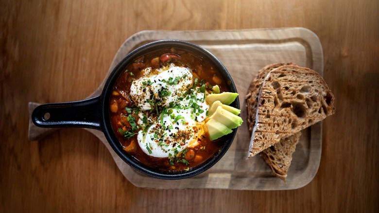 Tex Mex-style shakshuka in a cast iron pan on a wooden cutting board garnished with avocado and crusty bread