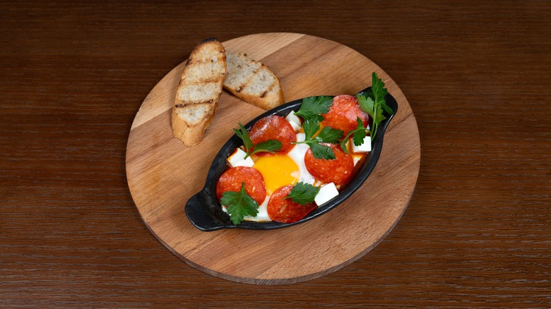 Oblong pan of shakshuka with feta on a wooden serving board with two slices of bread