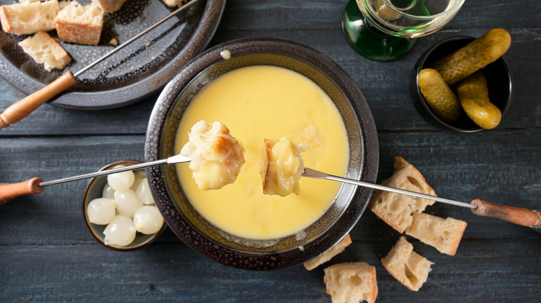 cheese fondue in a pot surrounded by cubes of bread, pickles, and pickled onions