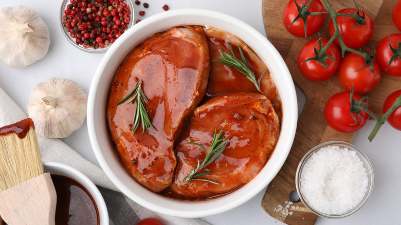 Meat marinating in a ceramic white bowl surrounded by salt, garlic, peppercorns, and tomatoes
