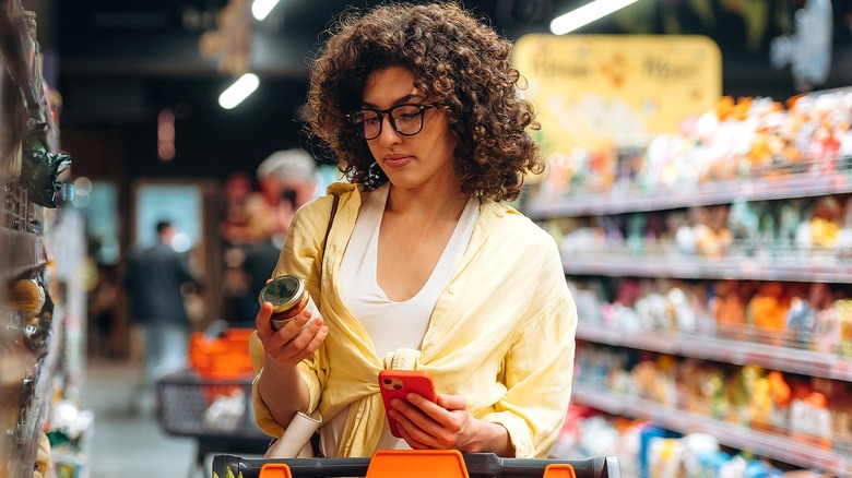 A woman reading the label of a can of food while holding her phone in a grocery store