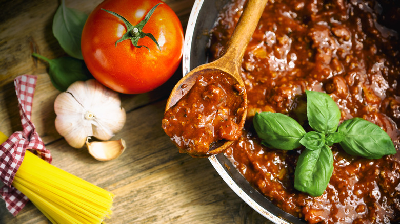 Bolognese sauce in a pot with fresh basil, a tomato, garlic cloves, and spaghetti