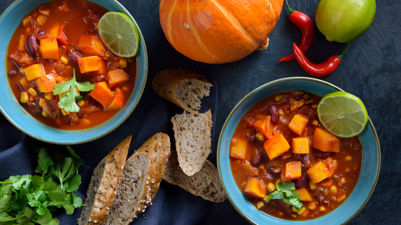 Two bowls of vegan chili on a blue background with slices of bread and a pumpkin