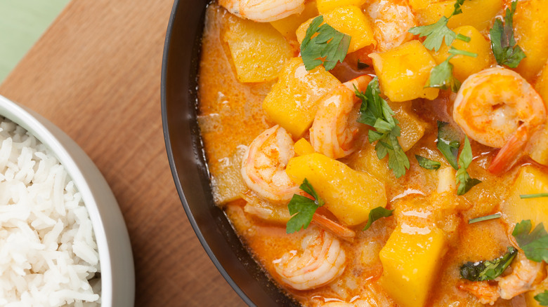 A close up picture of red curry with pumpkin and shrimp alongside a bowl of white rice