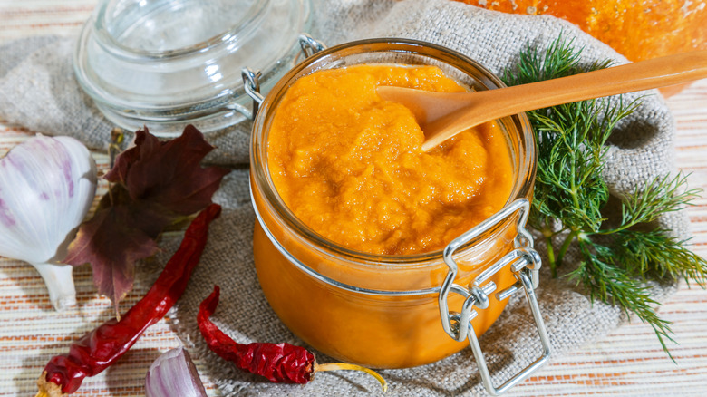 Pumpkin puree in a glass jar with a wooden spoon alongside herbs and spices on a gray napkin