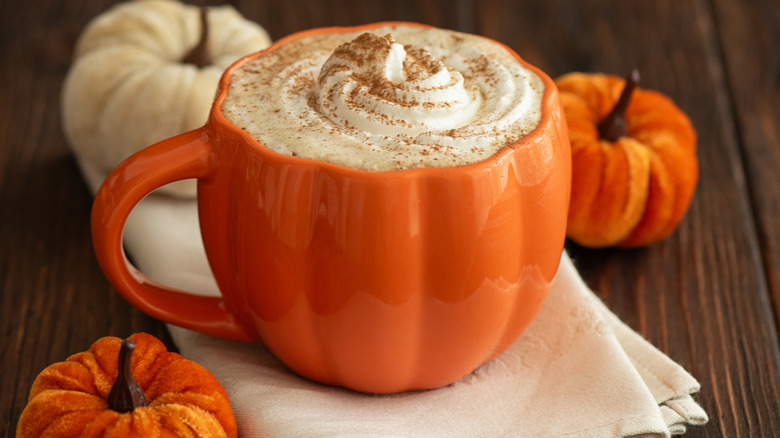 A pumpkin latte in a pumpkin-shaped cup on a white napkin surrounded by small pumpkins