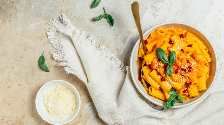 A bowl of rigatoni pasta in pumpkin sauce on a white napkin on a textured cream background