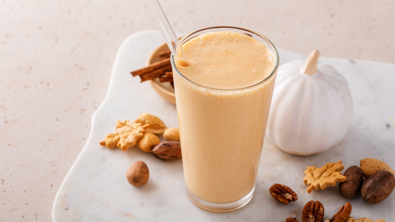 A pumpkin smoothie on a white marble board surrounded by a pumpkin, nuts, and leaf-shaped crackers