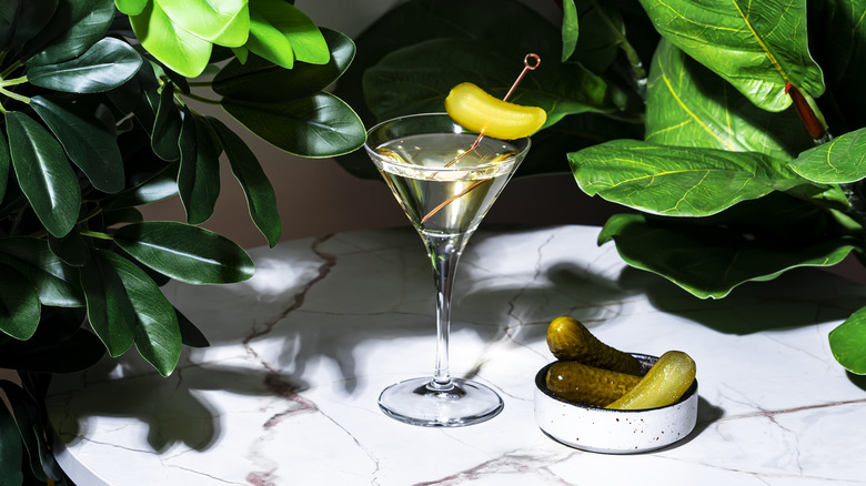 A martini with a pickle spear on a white countertop with green foliage