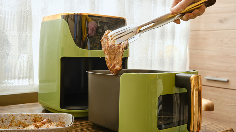 A hand holds tongs putting a piece of marinated meat to an air fryer.